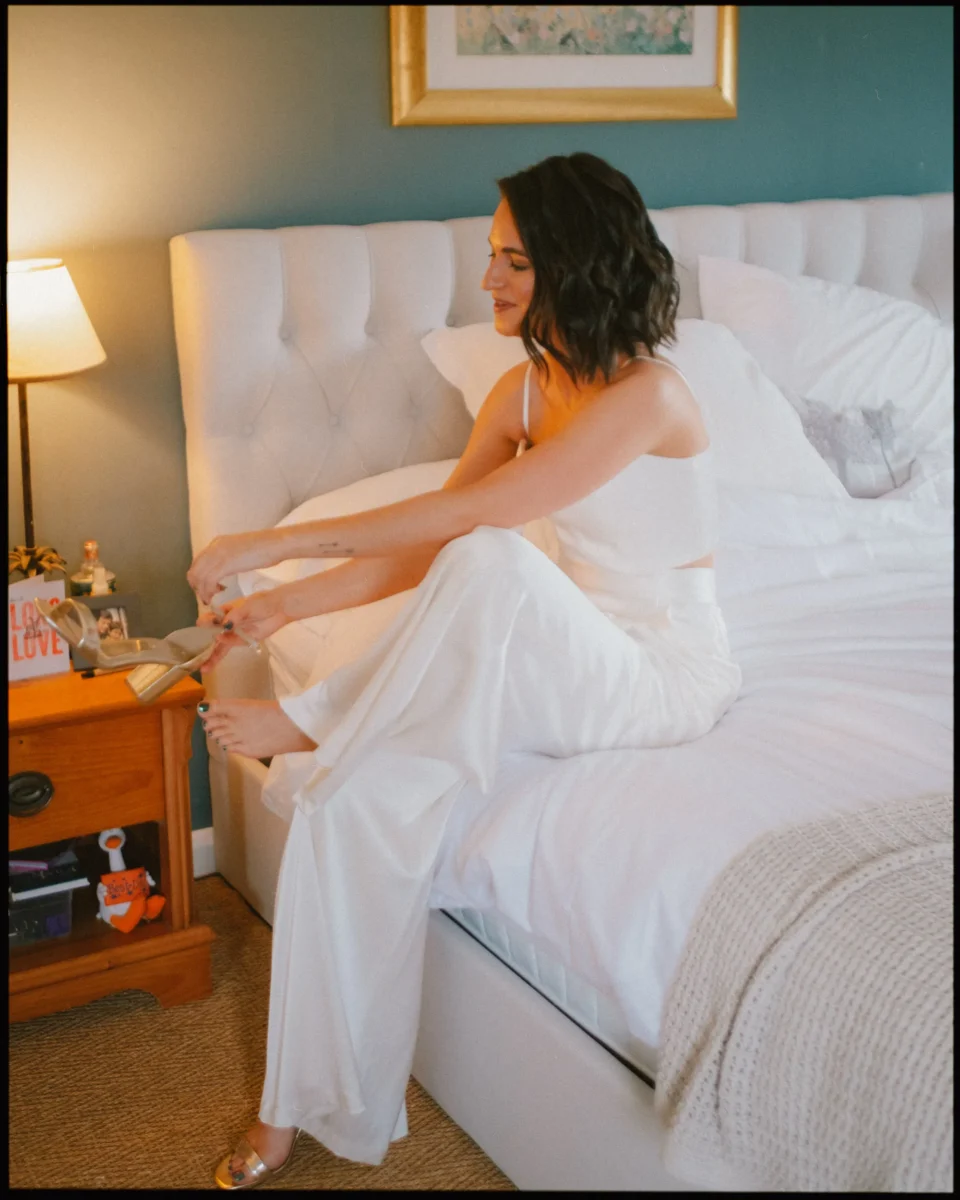 A woman with dark hair in a white outfit sits on a bed, smiling as she puts on gold shoes. The room has a teal wall, framed art, a bedside lamp, and a neatly made bed with white bedding.