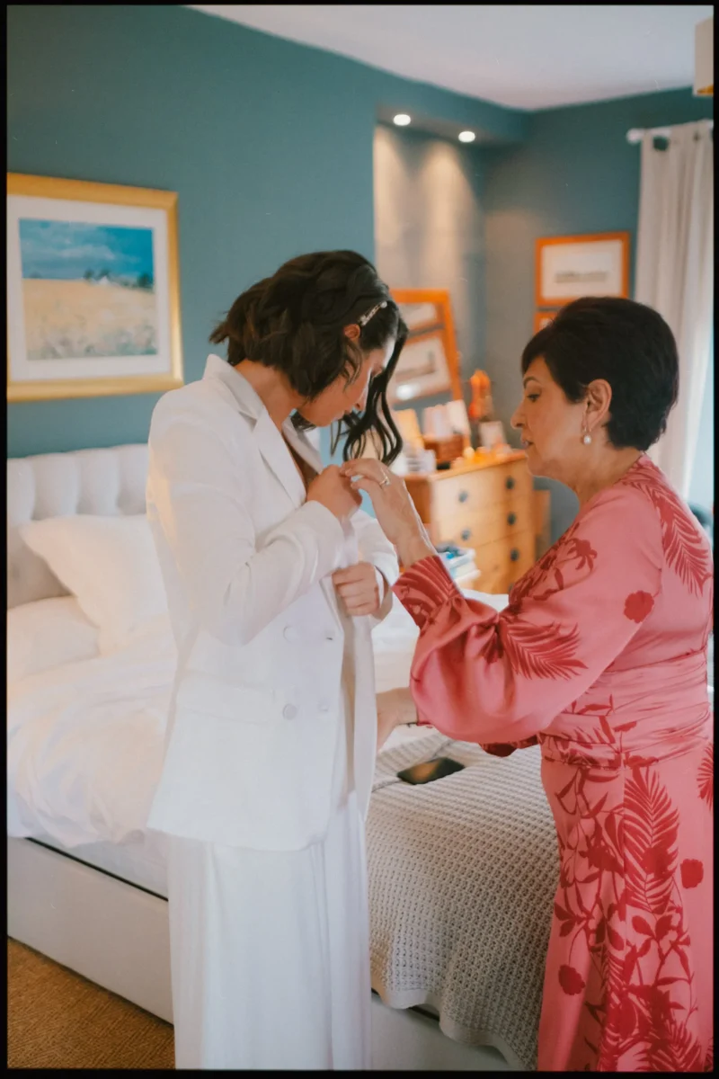 A woman in a white suit is being helped with her jacket by an older woman in a pink floral dress. They are standing next to a bed in a softly lit bedroom with teal walls and framed art.