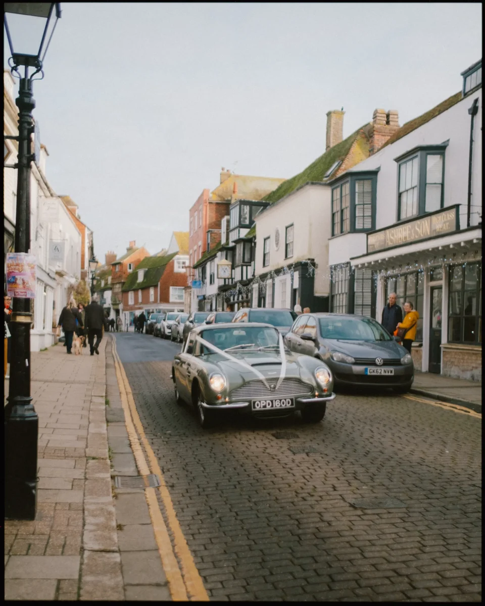 A vintage convertible decorated with white ribbon drives down a cobblestone street lined with historic buildings and shops. A few people walk on the sidewalk under a cloudy sky.