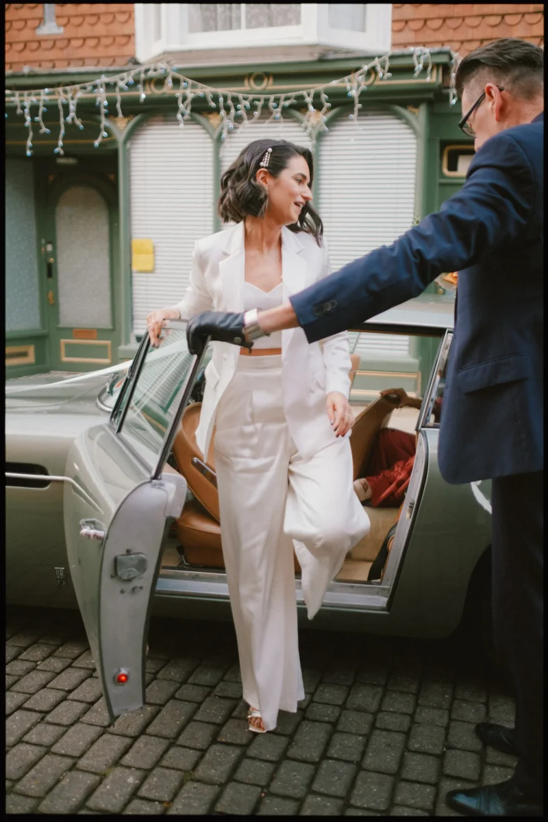A woman in a white suit steps out of a vintage car, smiling, while a man in a dark suit and gloves holds the door open for her on a cobblestone street, with festive lights in the background.