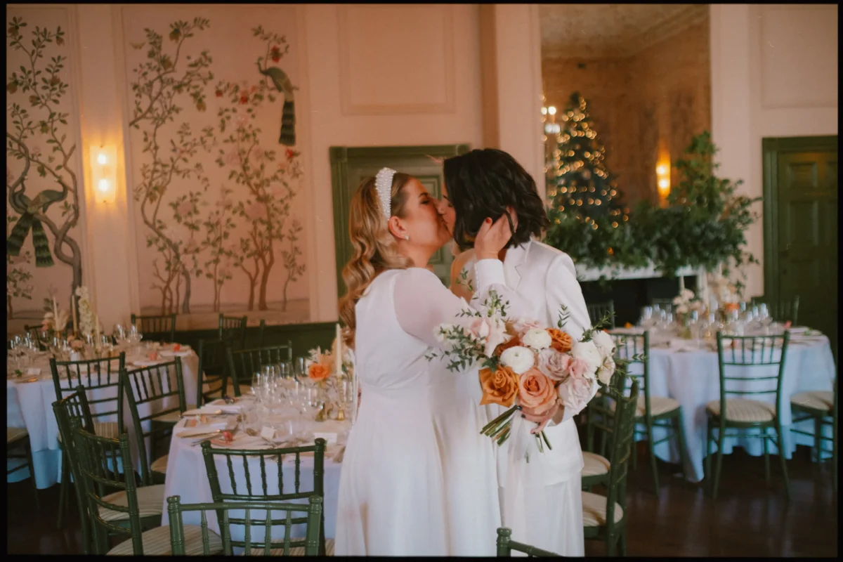 Two women in white wedding attire share a kiss in an elegantly decorated reception room with round tables, floral centerpieces, and a bouquet of peach and white flowers.