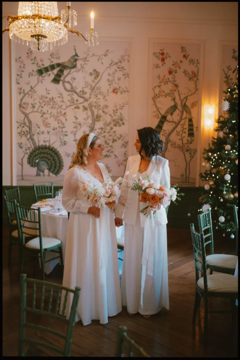 Two brides in white outfits stand holding bouquets, smiling at each other in an elegant room decorated with floral wall art, green chairs, a chandelier, and a Christmas tree with white lights and ornaments.