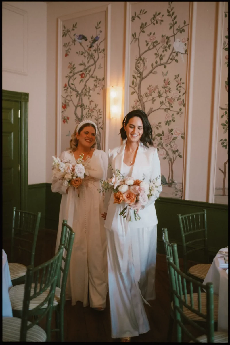 Two women in white wedding outfits, one in a dress and the other in a suit, walk down an aisle holding bouquets. They smile happily in an elegantly decorated room with floral wall panels and green chairs.