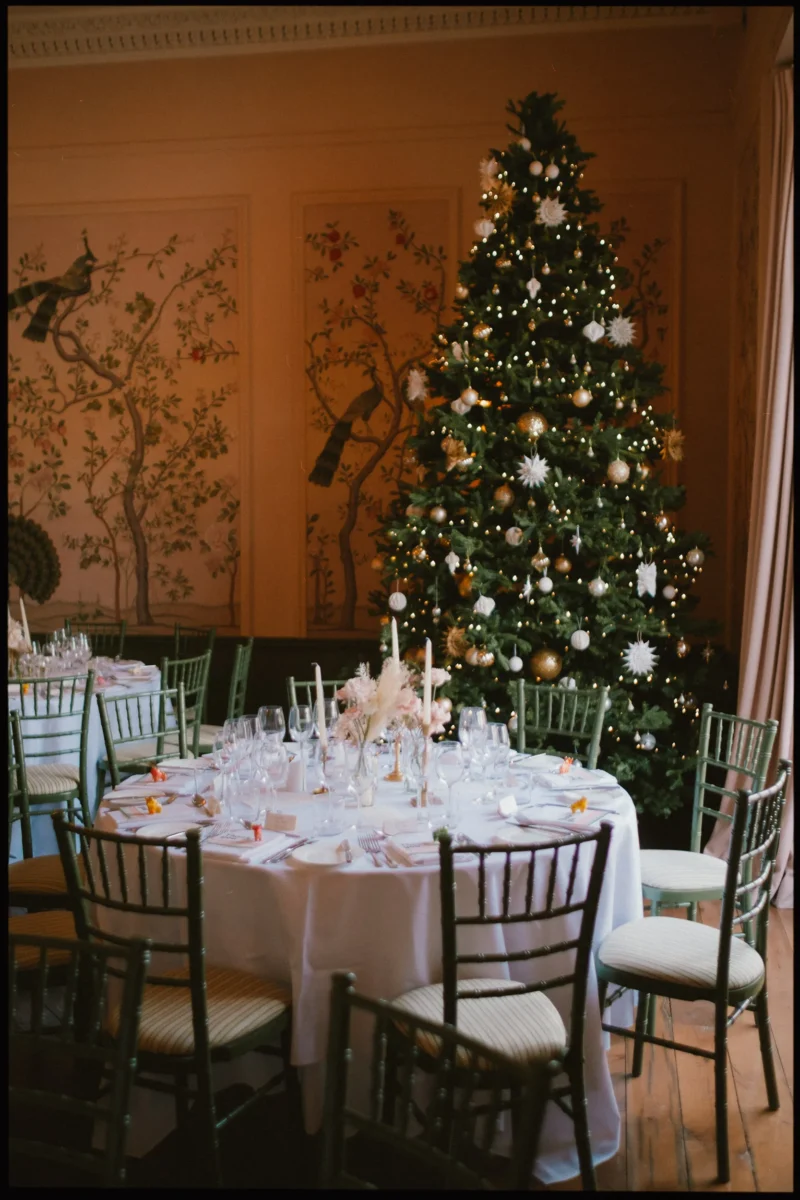 A round table set for a formal meal with glassware and candles, surrounded by green chairs, sits near a decorated Christmas tree with gold and white ornaments in an elegantly painted room.