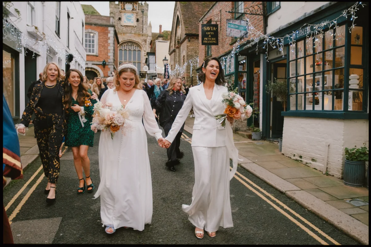 Two brides in white outfits walk hand in hand down a charming street, smiling and holding bouquets, followed by joyful friends. The street is lined with shops decorated with string lights.