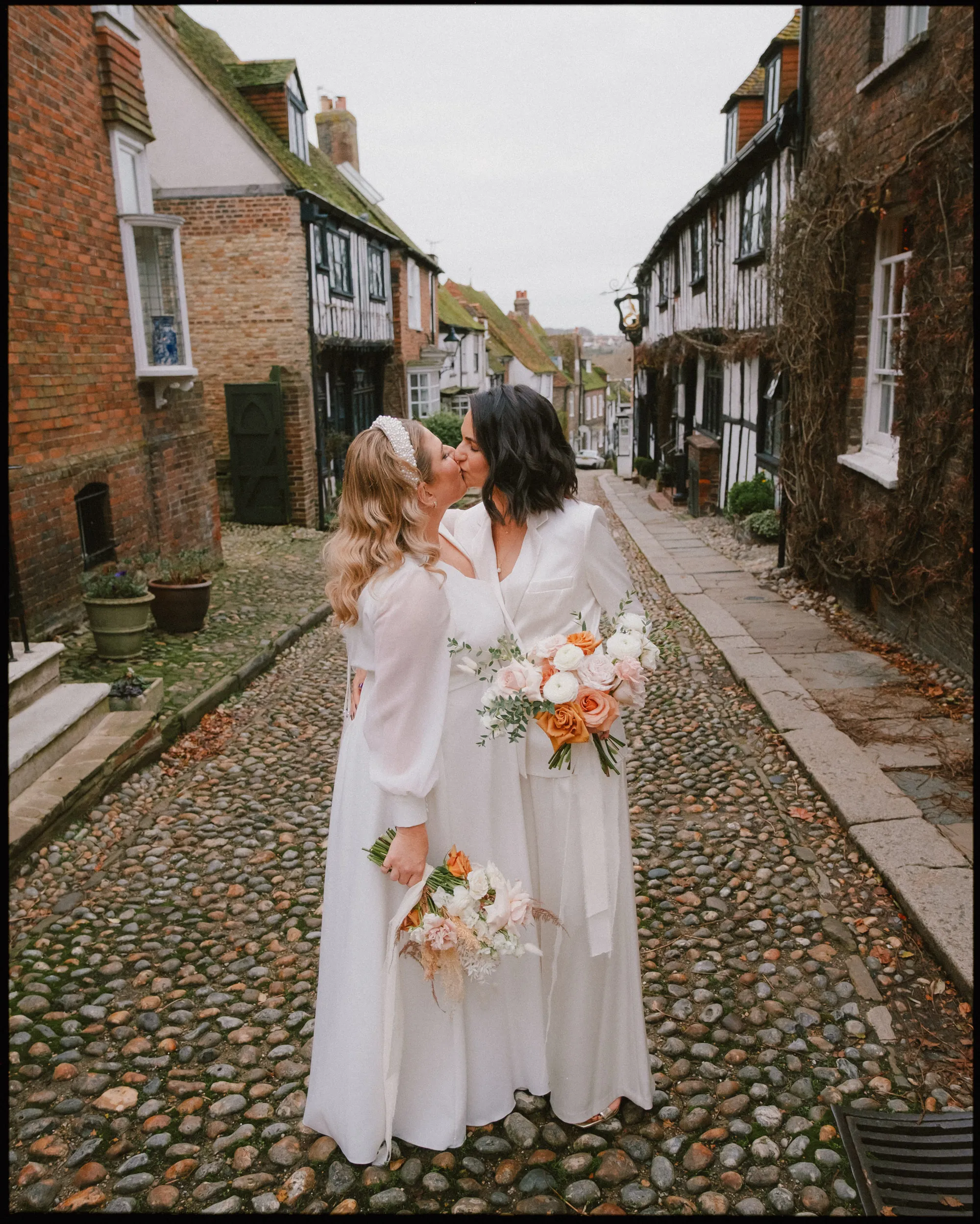 Two brides in white dresses and holding bouquets stand close together, sharing a kiss on a cobblestone street lined with old buildings, creating a romantic and joyful wedding scene.