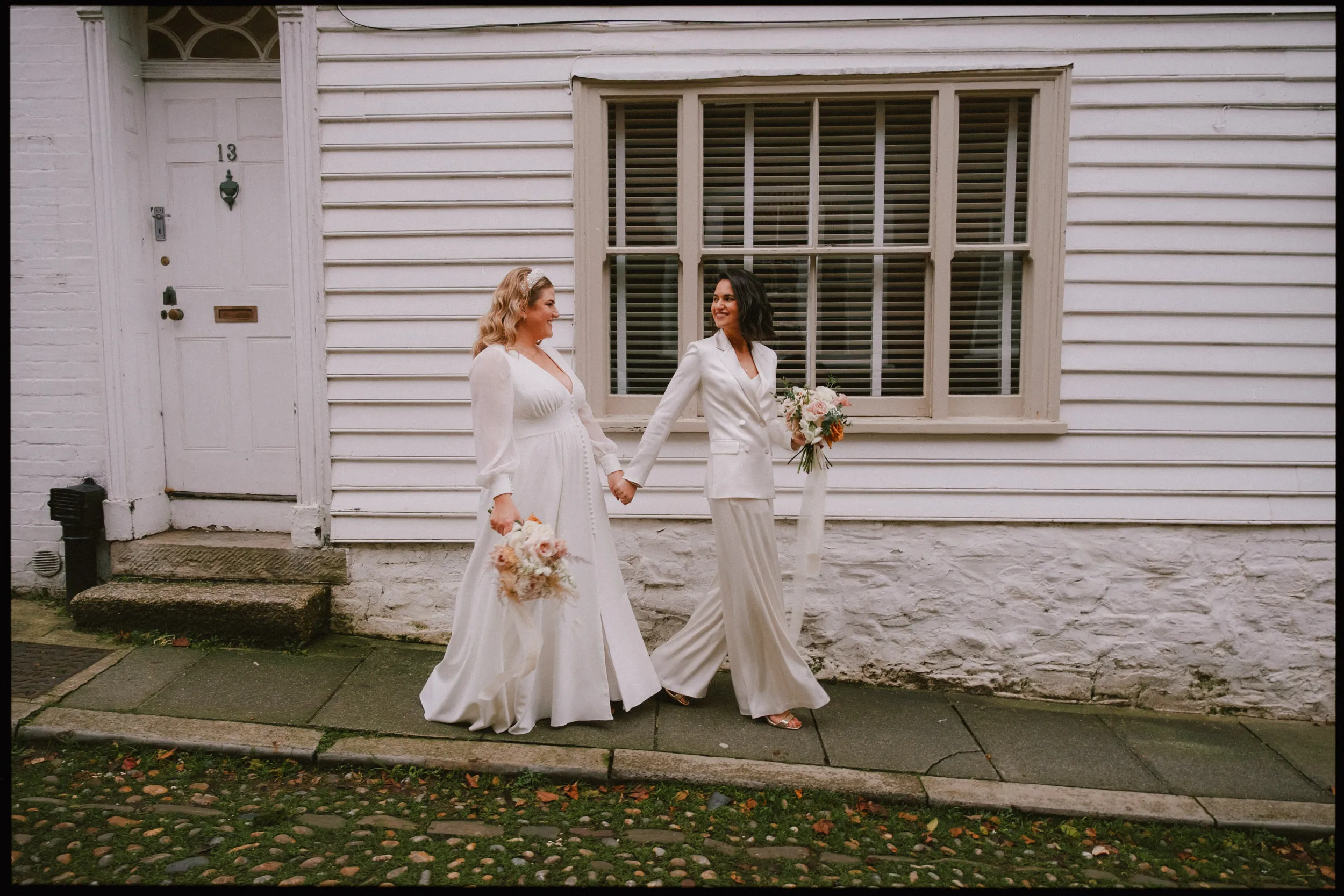 Two women wearing white outfits, holding hands and bouquets, walk together smiling along a cobblestone street in front of a white wooden building with a door and window.