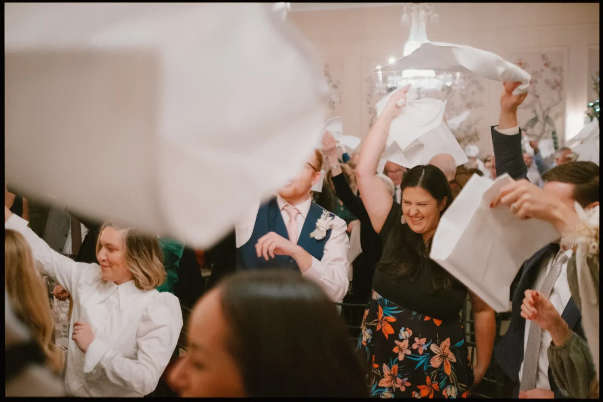 A group of people joyfully wave napkins in the air at an indoor celebration, smiling and wearing semi-formal attire. The scene is lively, with blurred motion and a chandelier visible in the background.
