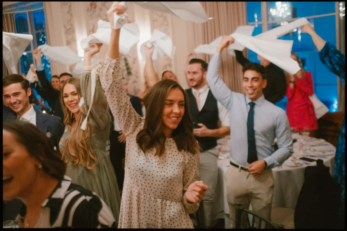 People at a formal event joyfully wave white napkins in the air, smiling and celebrating in a warmly lit, elegant room with tables and chandeliers.
