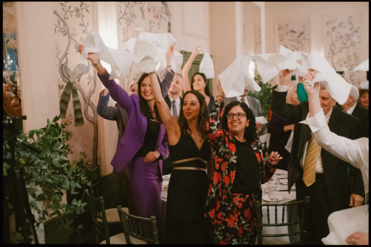 A group of well-dressed people stand around a dining table indoors, smiling and waving white napkins in the air in celebration. The room is decorated with floral wallpaper and greenery.