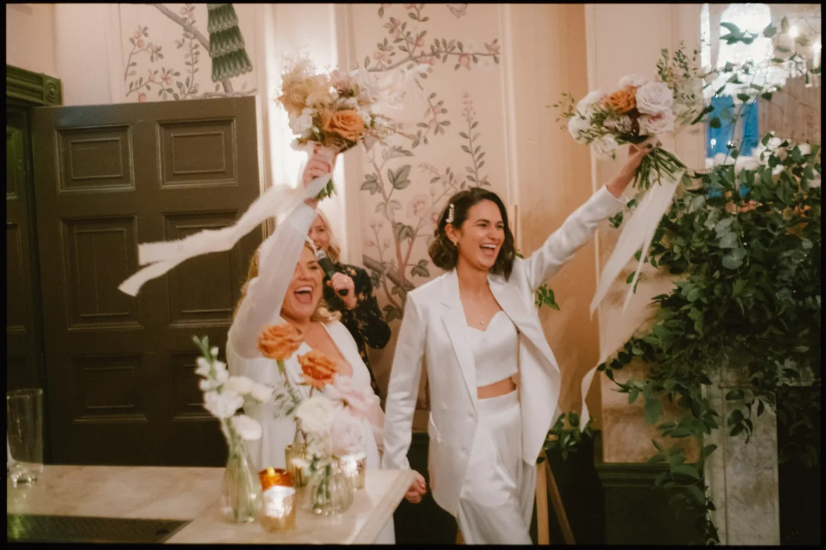 Two women, one in a white suit and the other in a white dress, smile joyfully and raise bouquets in the air while holding hands in a warmly decorated room with flowers and candles.