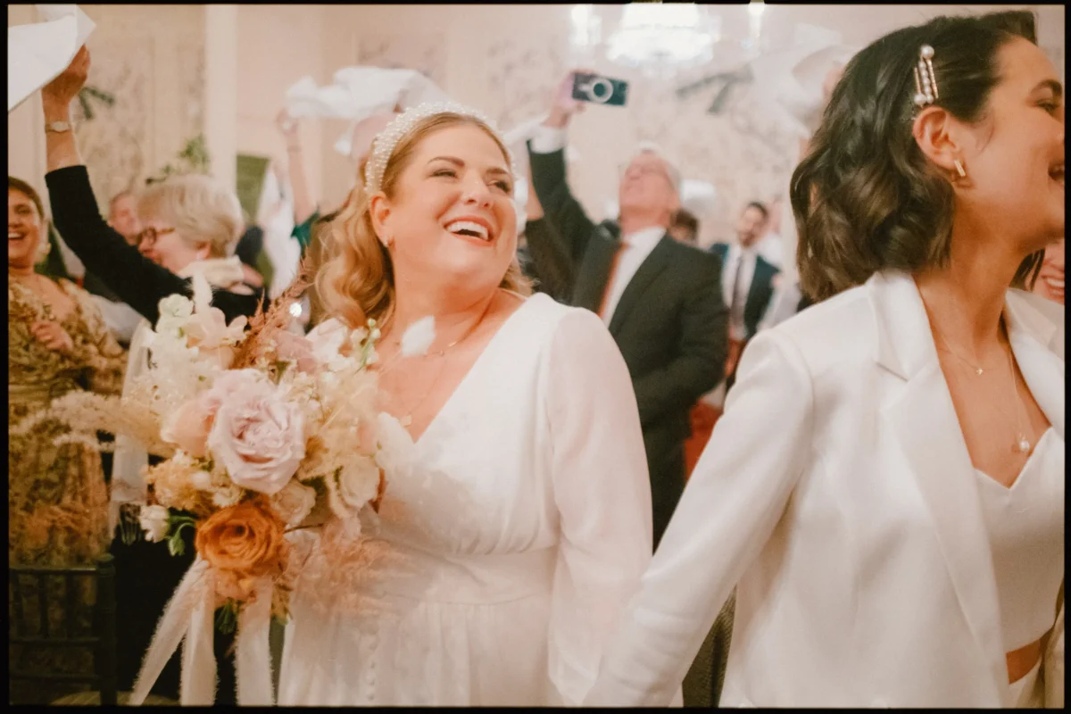 Two women in white outfits smile and celebrate at a wedding ceremony. One holds a bouquet of pink and beige flowers. Guests in the background wave napkins and take photos, creating a joyful atmosphere.