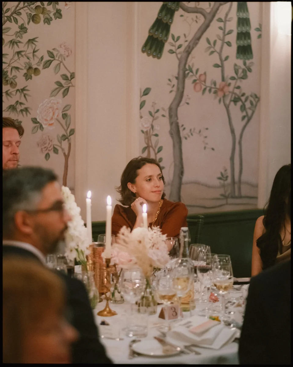 A woman in a red top sits at an elegantly set dinner table with candles, flowers, and glassware, surrounded by other guests. The background features floral and bird-themed wall art.