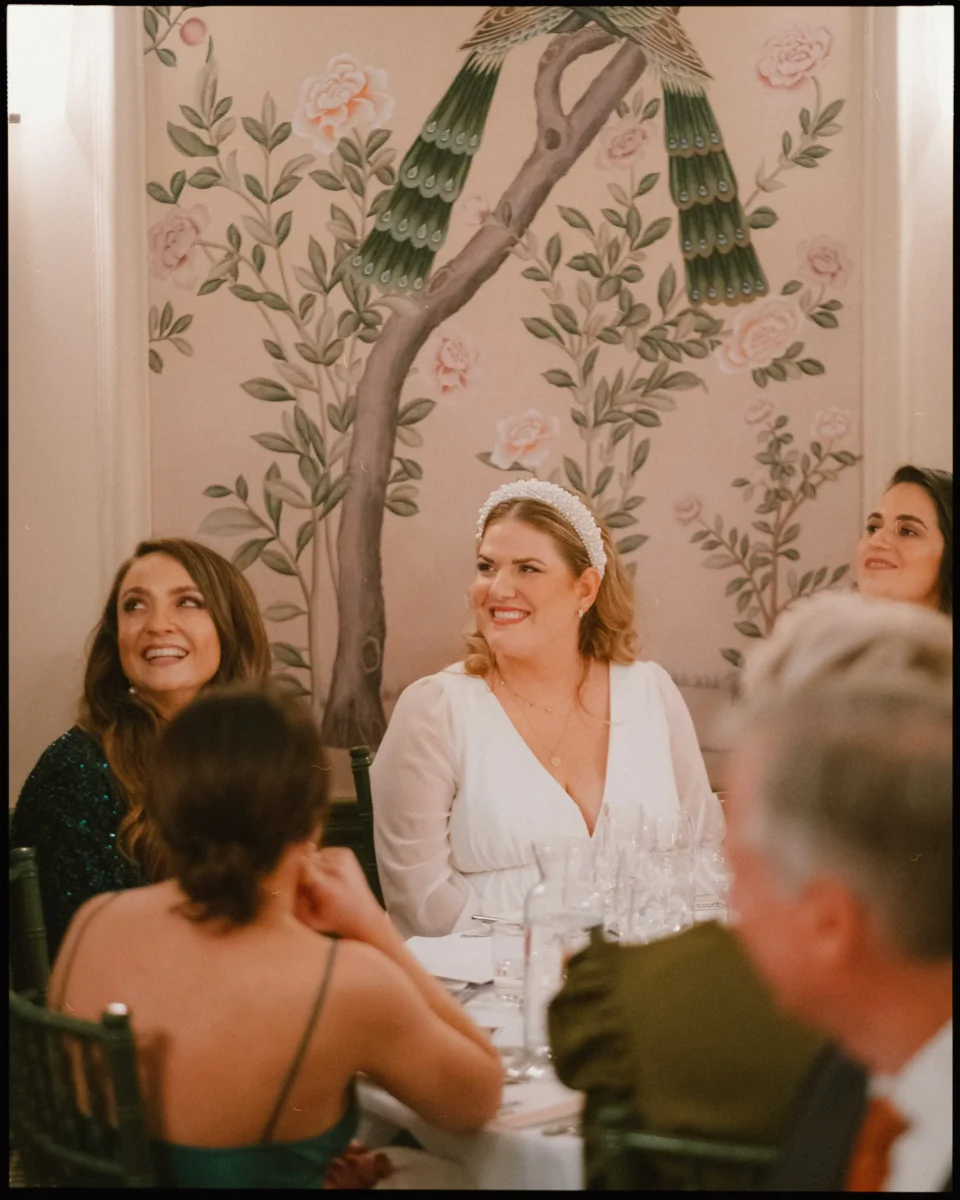 Four women in formal attire sit smiling at a decorated round table, with glassware in front of them. Behind them is a wall mural of a large peacock and pink flowers. The scene appears joyful and celebratory.