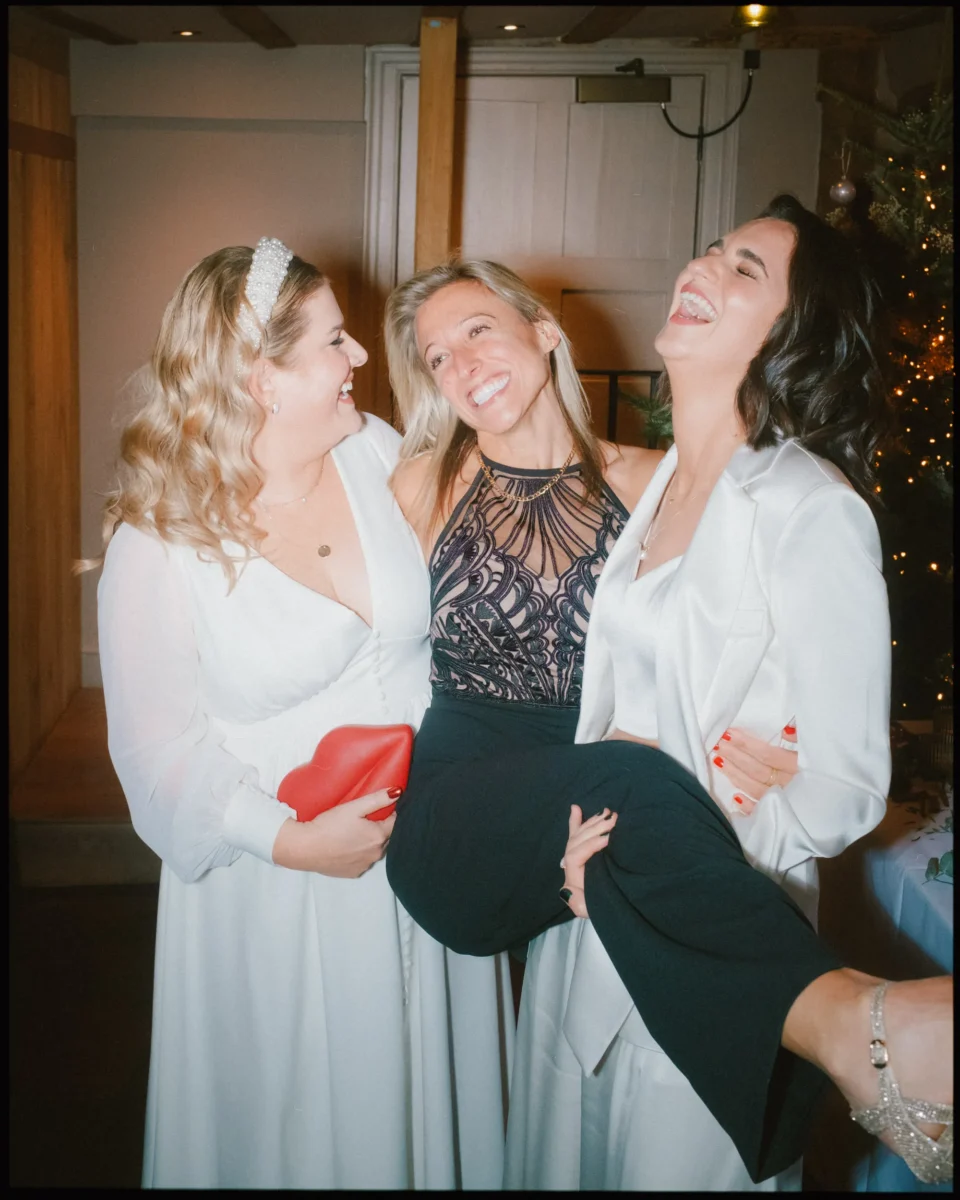 Three women smiling and laughing at a formal event; two women in white outfits hold a woman in black pants and a patterned top, who is being lifted and is laughing joyfully. A decorated Christmas tree is in the background.