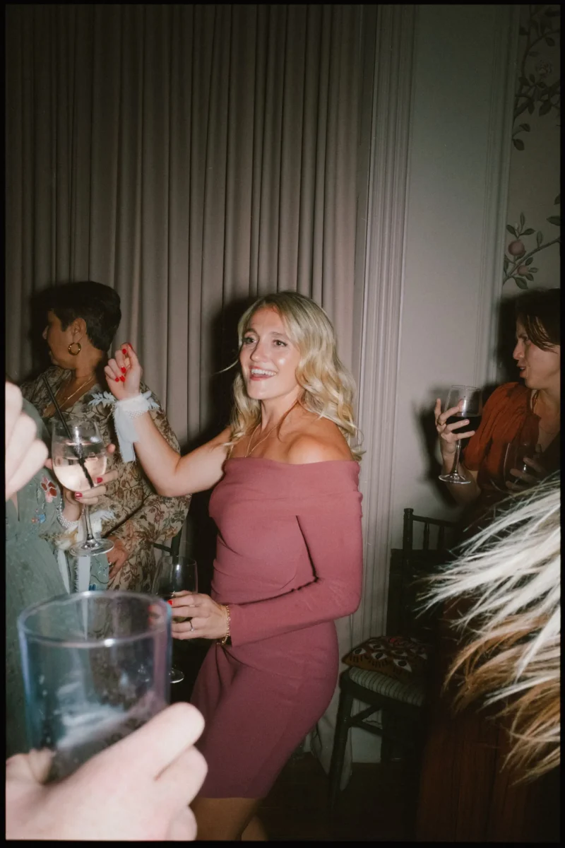 A woman in a pink off-the-shoulder dress smiles and dances at a party, holding a glass of wine. Other guests around her also hold drinks and appear to be enjoying themselves.