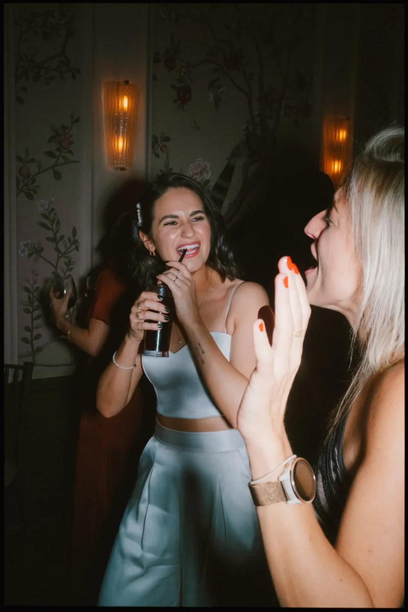 Two women laugh and smile while enjoying drinks at a party. One wears a white crop top and pants, holding a can, while the other has painted nails and gestures animatedly. Warm lighting and floral wallpaper are visible in the background.