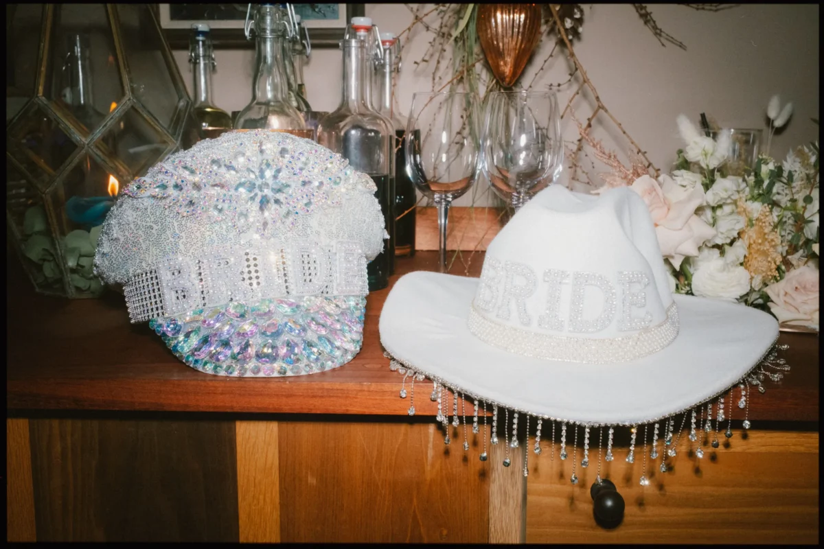 Two decorative bridal hats sit on a wooden surface: one sparkly and jeweled with BRIDE in rhinestones, the other a white cowboy hat with pearl fringe and BRIDE in glitter. Bottles, glasses, and flowers are in the background.