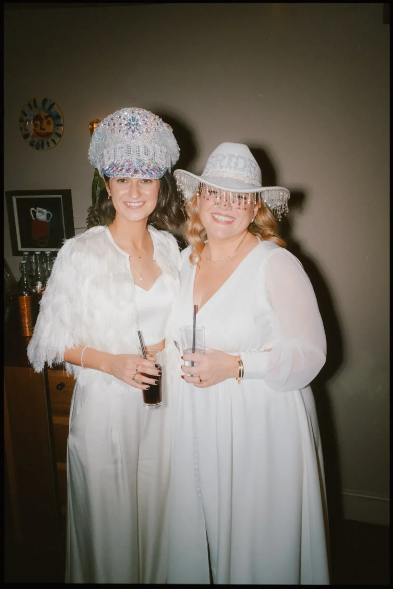 Two women dressed in white outfits and holding drinks smile at the camera. They both wear decorative hats; one hat reads BRIDE. The background includes a clock, artwork, and a drinks station.