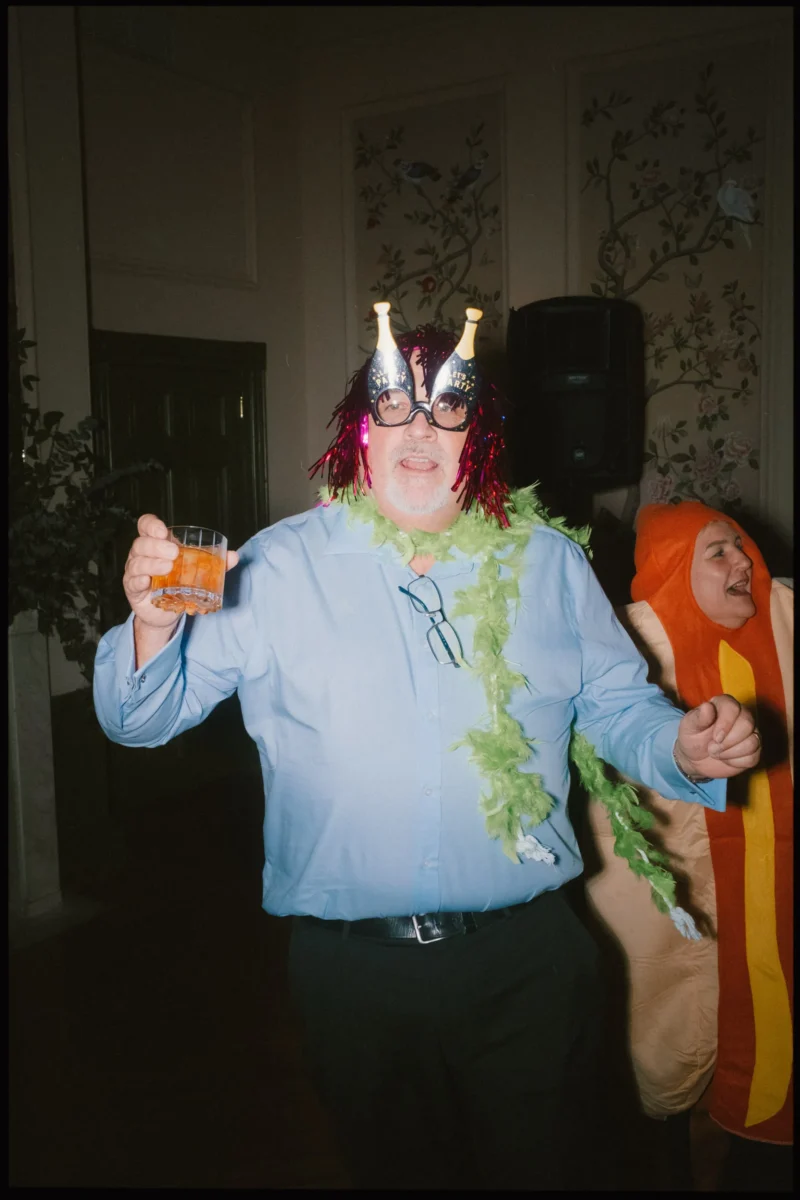 A man wearing novelty glasses with champagne bottles, a red tinsel wig, a green feather boa, and holding a drink stands next to someone in a hot dog costume at a costume party.
