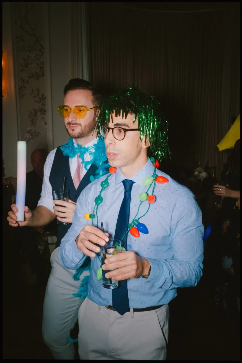 Two men at a party wear festive accessories: one has yellow sunglasses and a blue feather boa, the other has a green tinsel wig and Christmas lights around his neck. Both hold drinks and glow sticks, enjoying the event.