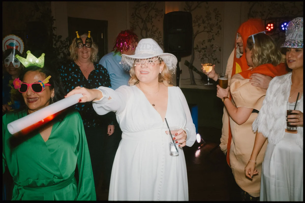 A woman in a white dress and bride hat points a glowing stick while others in costumes—such as a green crown, bug antennae, and food outfit—celebrate and smile at a lively party.