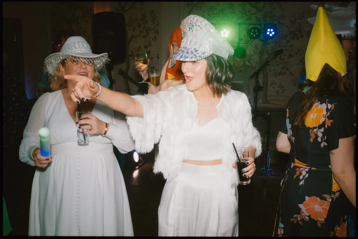 A woman in a white outfit and sparkling hat points and smiles at a lively party, surrounded by other guests in colorful hats and holding drinks, under festive lighting.