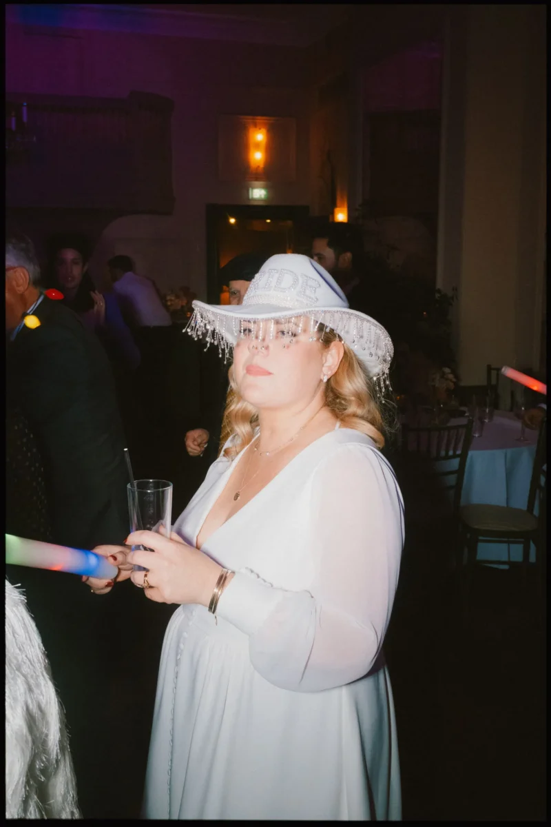 A woman in a white dress and a BRIDE hat with fringe holds a drink at a party, surrounded by colorful lights and other guests in a dimly lit room.