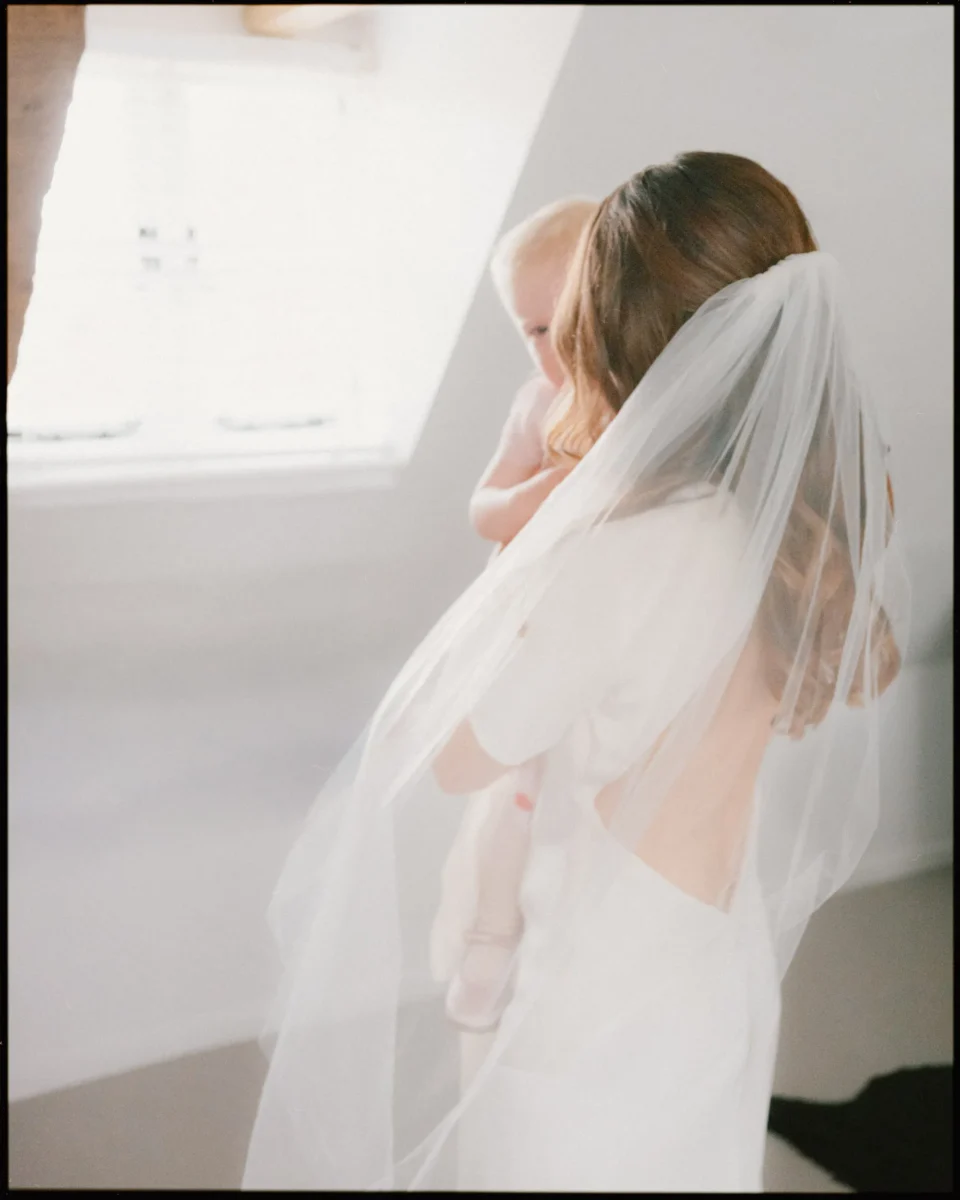 A bride with long brown hair in a white dress and veil holds a baby in a softly lit room with a window in the background.