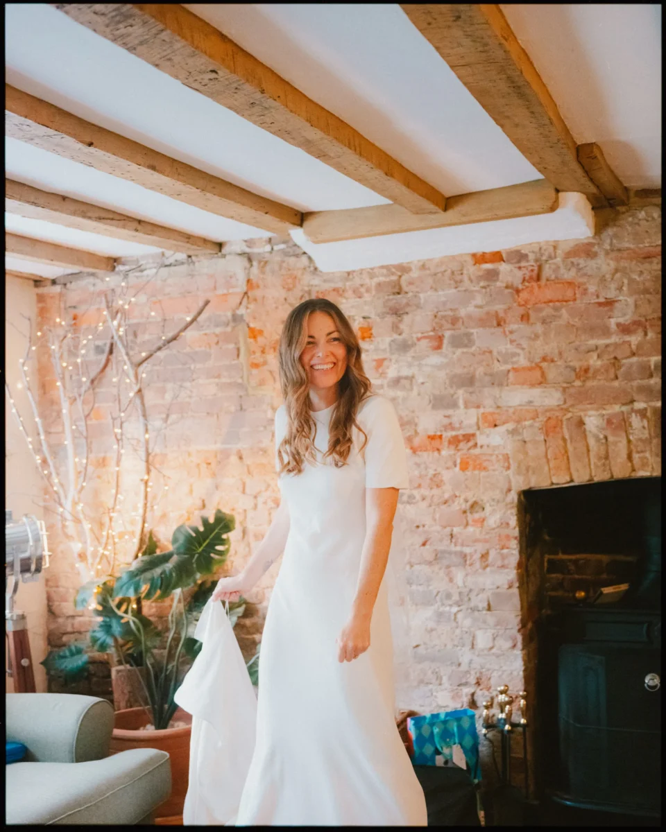 A woman in a white dress stands indoors, smiling joyfully. She is in a cozy room with exposed brick walls, wooden ceiling beams, potted plants, and string lights creating a warm, inviting atmosphere.