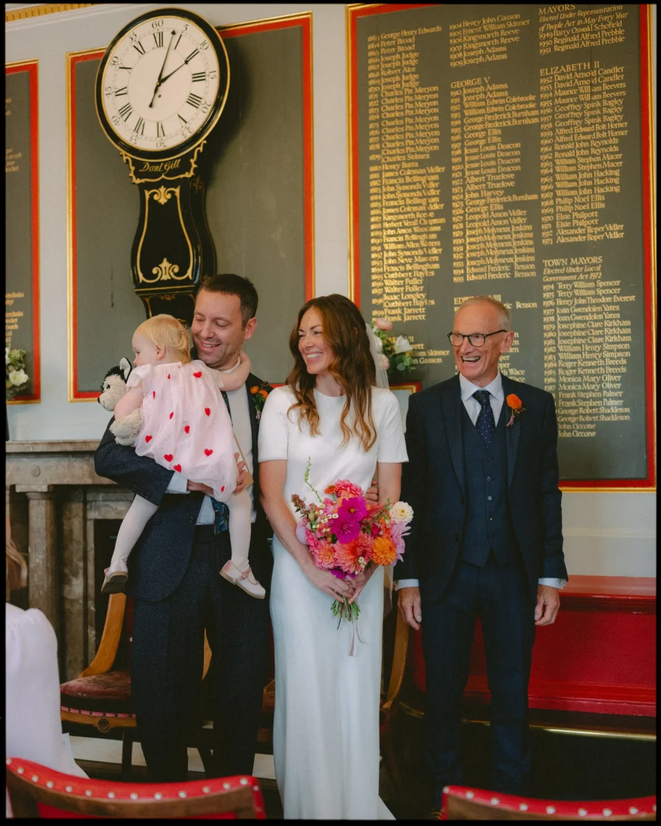 A couple stands smiling with a toddler in the mans arms and an older man beside them. The woman holds a bright bouquet. Behind them is a large clock and a wall with gold names listed on a board.