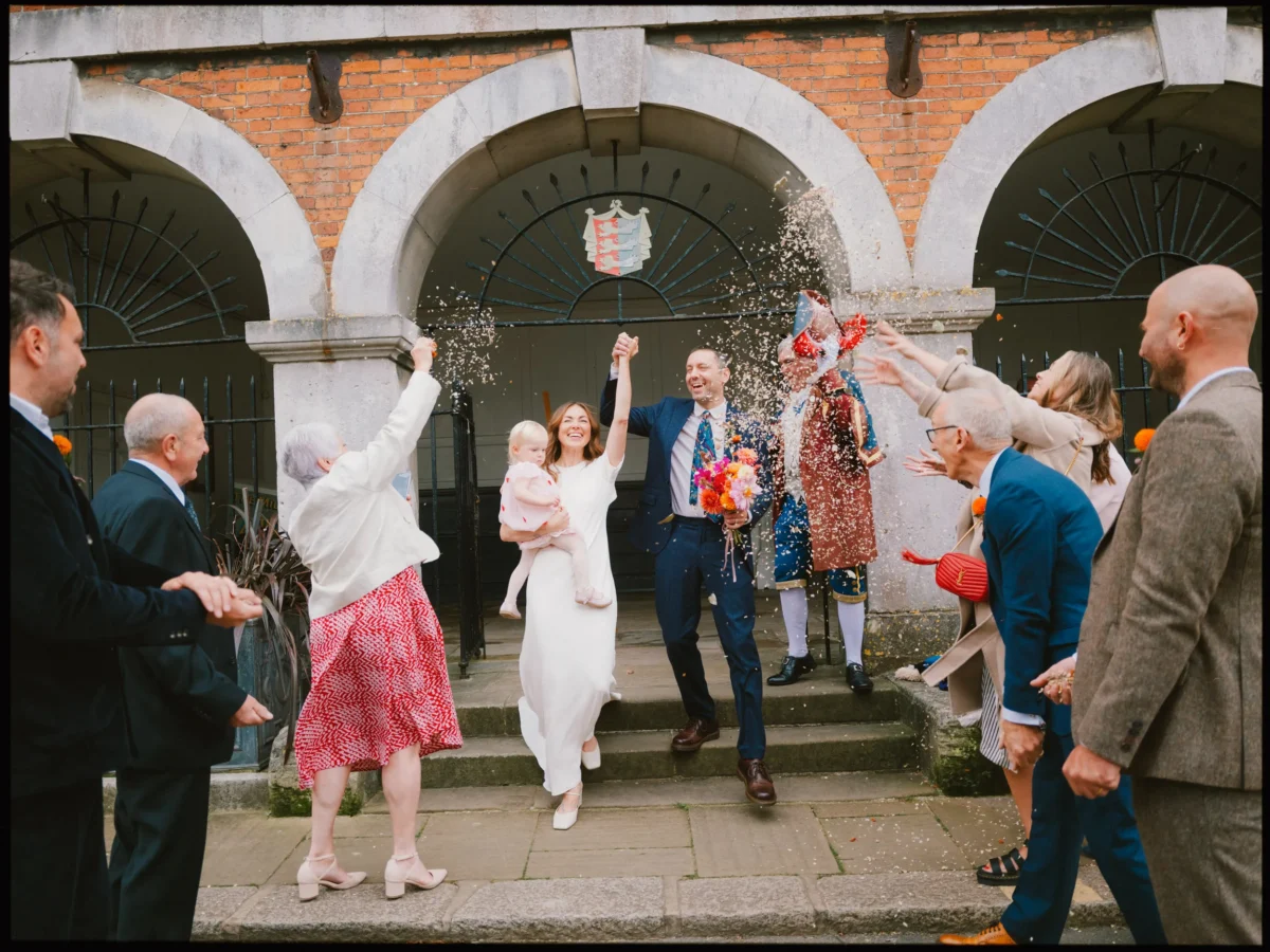 A newlywed couple, with the bride holding a young child, walk hand-in-hand under an archway as smiling guests celebrate and throw confetti at them. The group appears joyful and dressed in colorful attire.