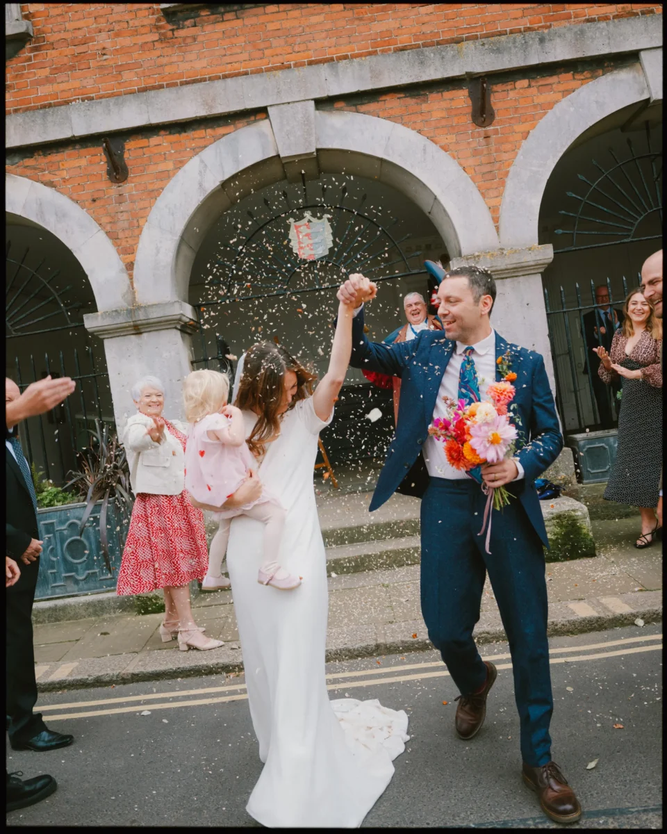A newlywed couple celebrates outside a building. The bride holds a child and lifts her hand with the groom as confetti falls around them. The groom holds a vibrant bouquet. Smiling guests clap and cheer in the background.