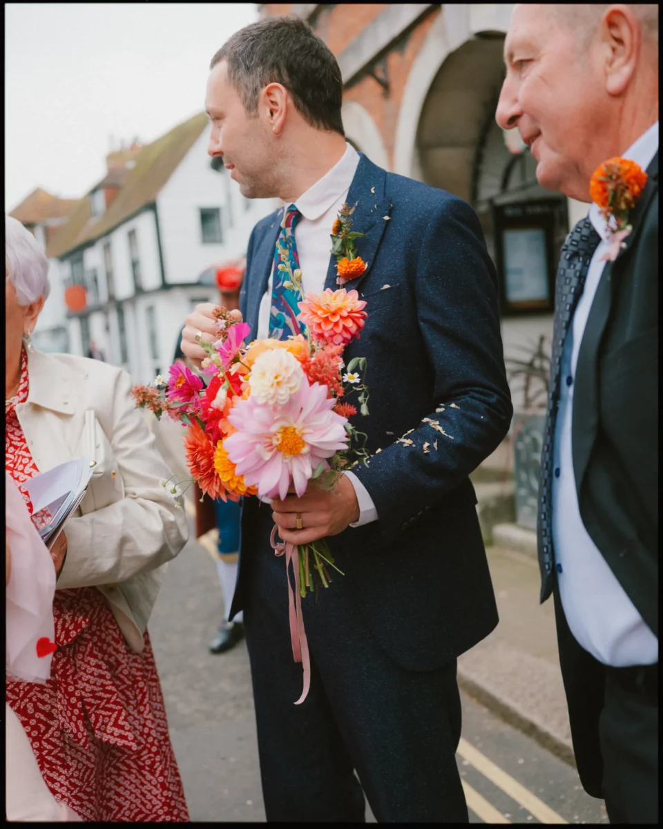 A man in a dark suit holding a colorful bouquet of flowers stands next to two older adults on a street. Flower petals are scattered on his suit, suggesting a festive or wedding event.