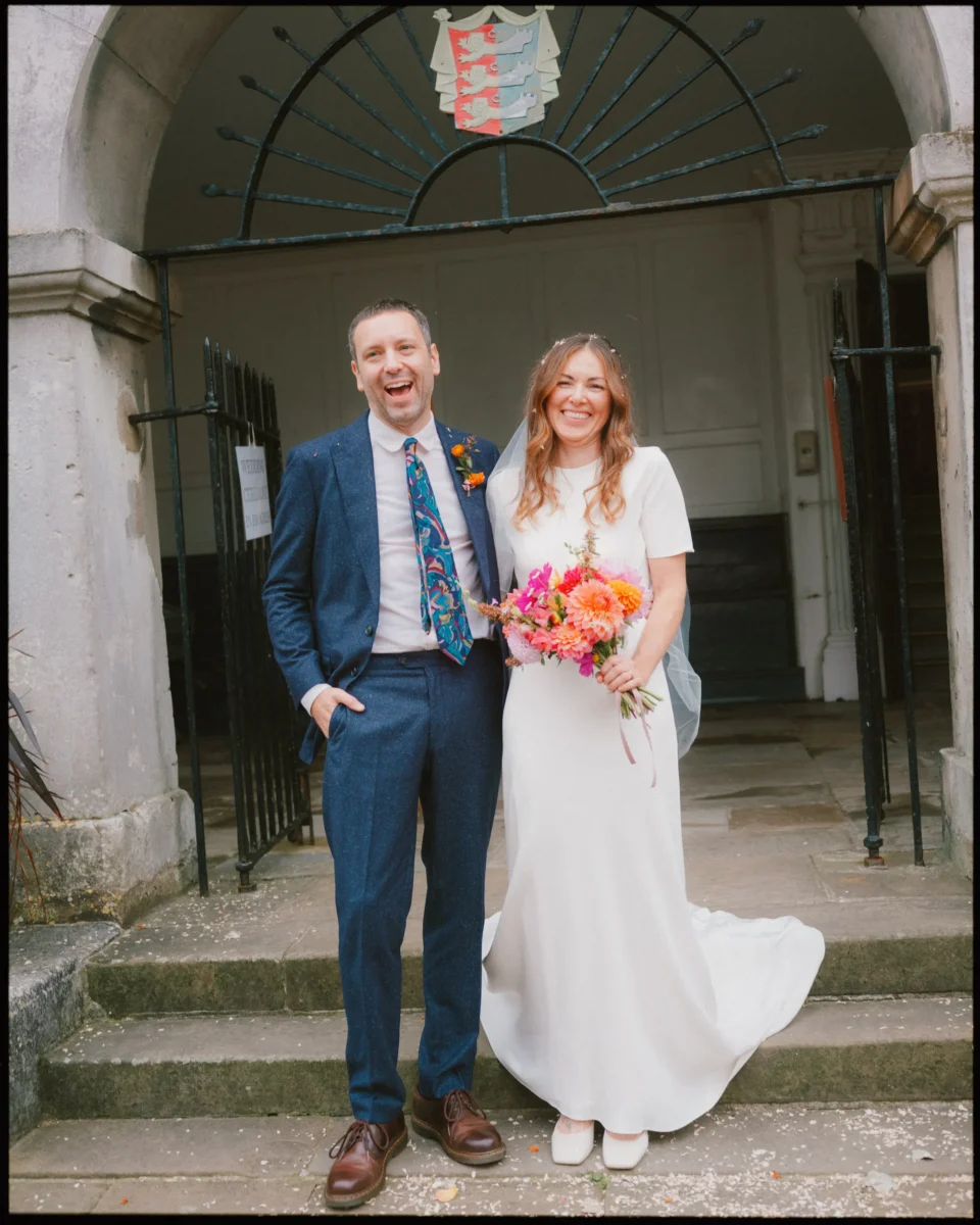 A smiling couple stands on stone steps outside a building with an arched entrance. The man wears a blue suit and patterned tie; the woman wears a white dress and holds a colorful bouquet. They appear happy, likely at a wedding.