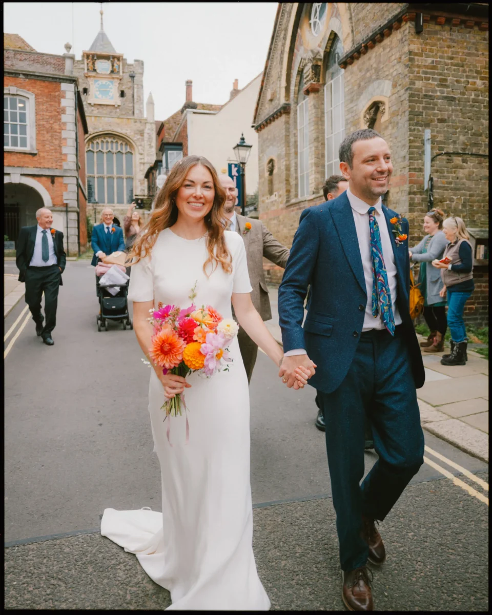 A smiling bride in a white dress holding a colorful bouquet walks hand-in-hand with a groom in a blue suit and patterned tie down a street, with friends and family following behind them.