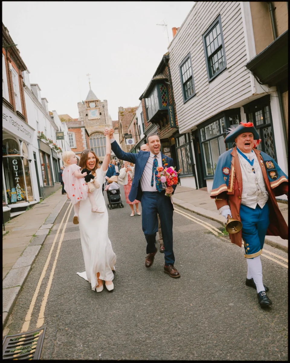 A joyful bride and groom walk down a street, the bride holding a child and the groom holding colorful flowers. A town crier leads the way, ringing a bell, as people in the background watch and take photos.