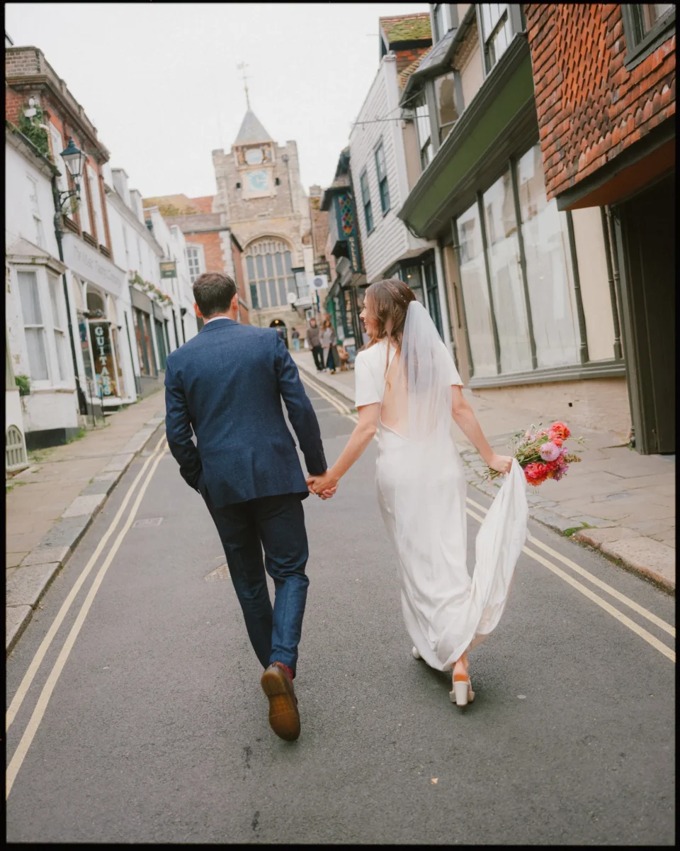 A bride and groom walk hand in hand down a cobblestone street lined with old buildings. The bride holds her dress and bouquet, while the groom is in a blue suit. They are seen from behind, heading toward a historic church.