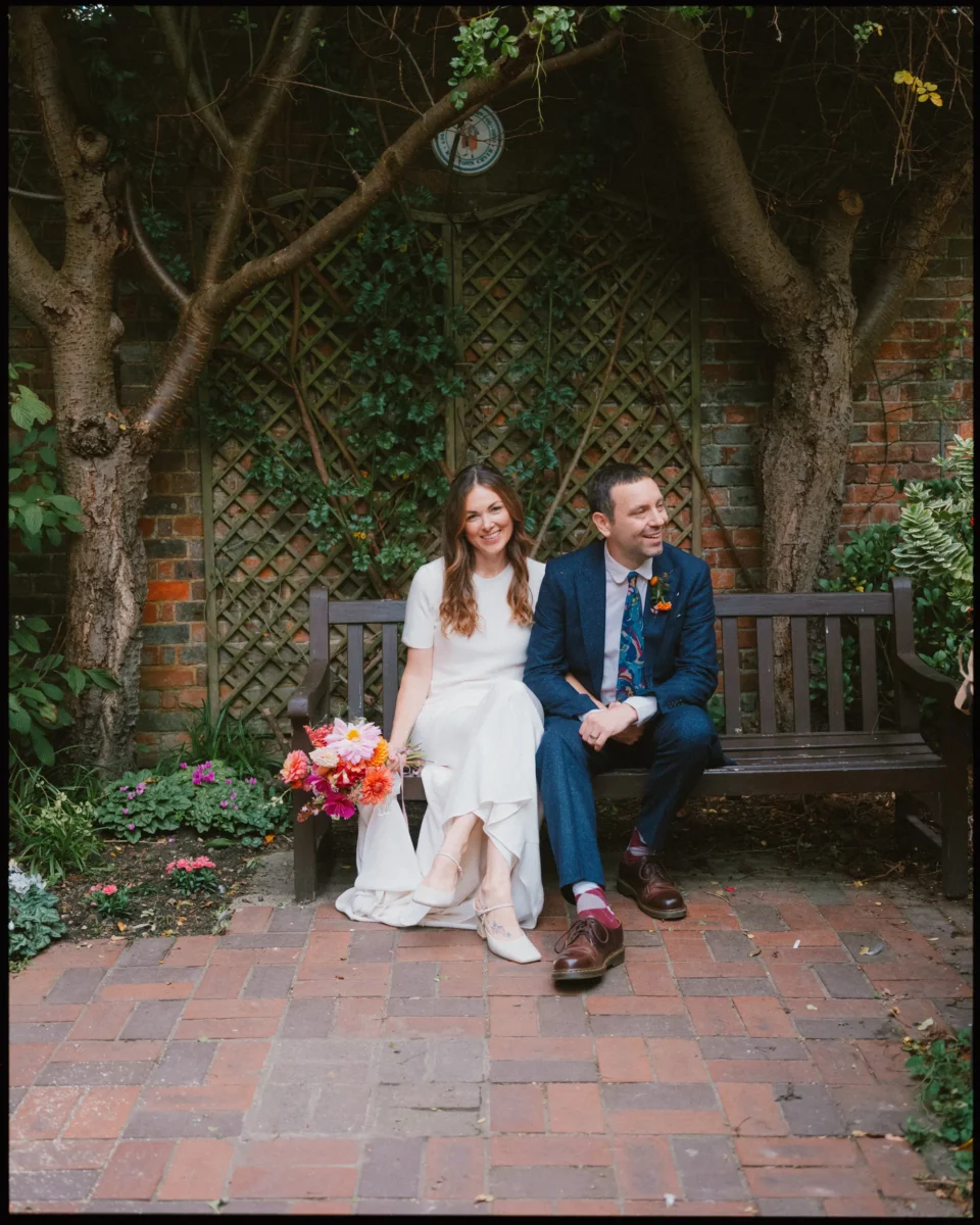 A bride in a white dress and groom in a blue suit sit smiling on a wooden bench in a garden, surrounded by greenery and flowers, with a colorful bouquet resting on the brides lap.