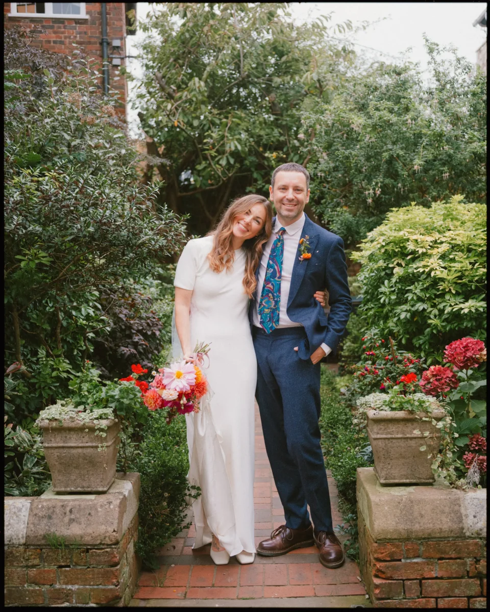 A smiling couple stands together in a lush garden; the woman wears a white dress and holds a bouquet of flowers, while the man wears a blue suit and patterned tie. They stand on a brick path surrounded by greenery and red flowers.