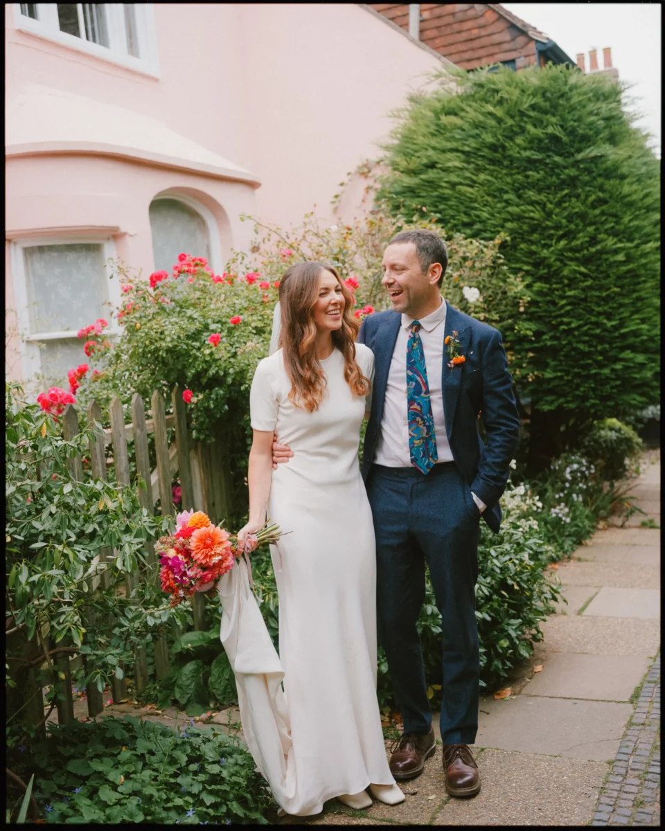 A bride in a white dress holding a colorful bouquet stands smiling next to a groom in a blue suit and patterned tie. They are outside by a garden fence with pink flowers and a pink house in the background.