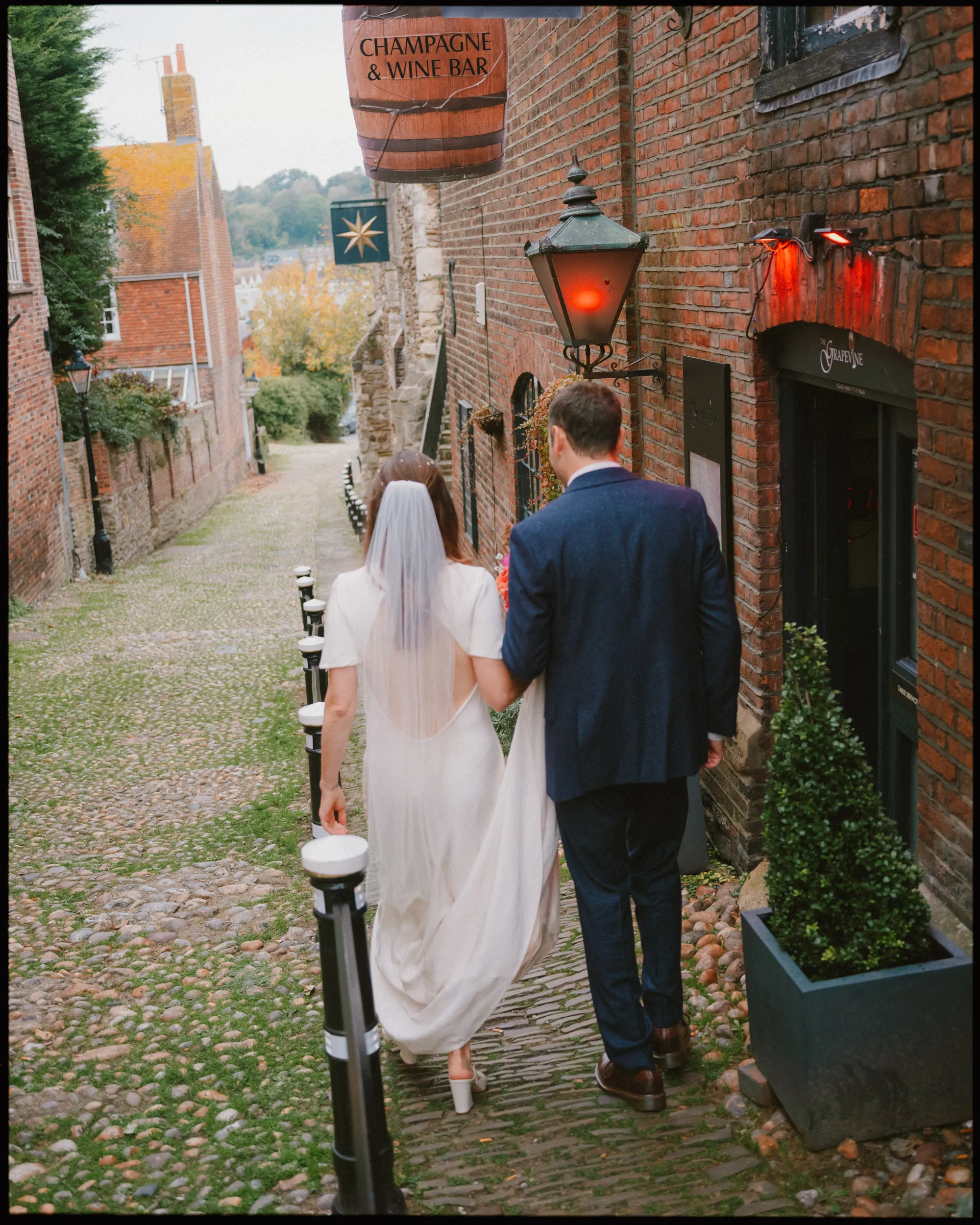 A bride and groom walk down a cobblestone street lined with brick buildings. The bride wears a white dress and veil, and the groom is in a blue suit. A champagne and wine bar sign hangs above them.