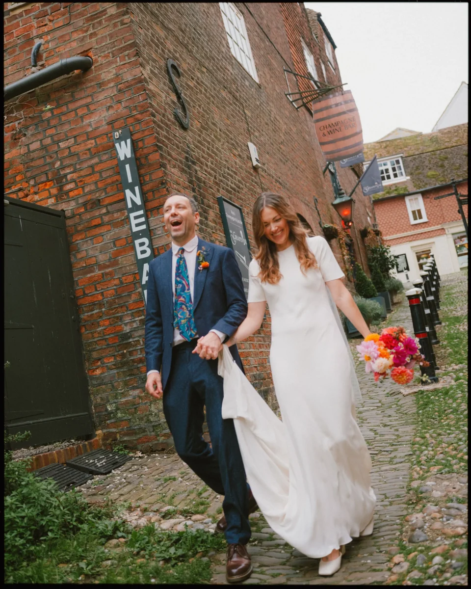 A smiling bride and groom walk hand in hand down a cobblestone street. The bride wears a white dress and holds a colorful bouquet, while the groom is in a blue suit with a patterned tie. They appear joyful beside a “WINE BAR” sign.