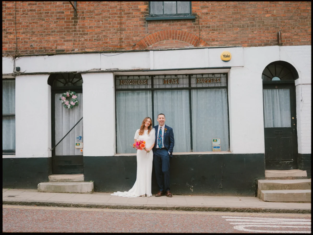 A bride in a white dress holding a bouquet and a groom in a suit smile while standing on a sidewalk in front of an old brick building with lace-curtained windows and a sign reading Luncheons Teas Suppers.