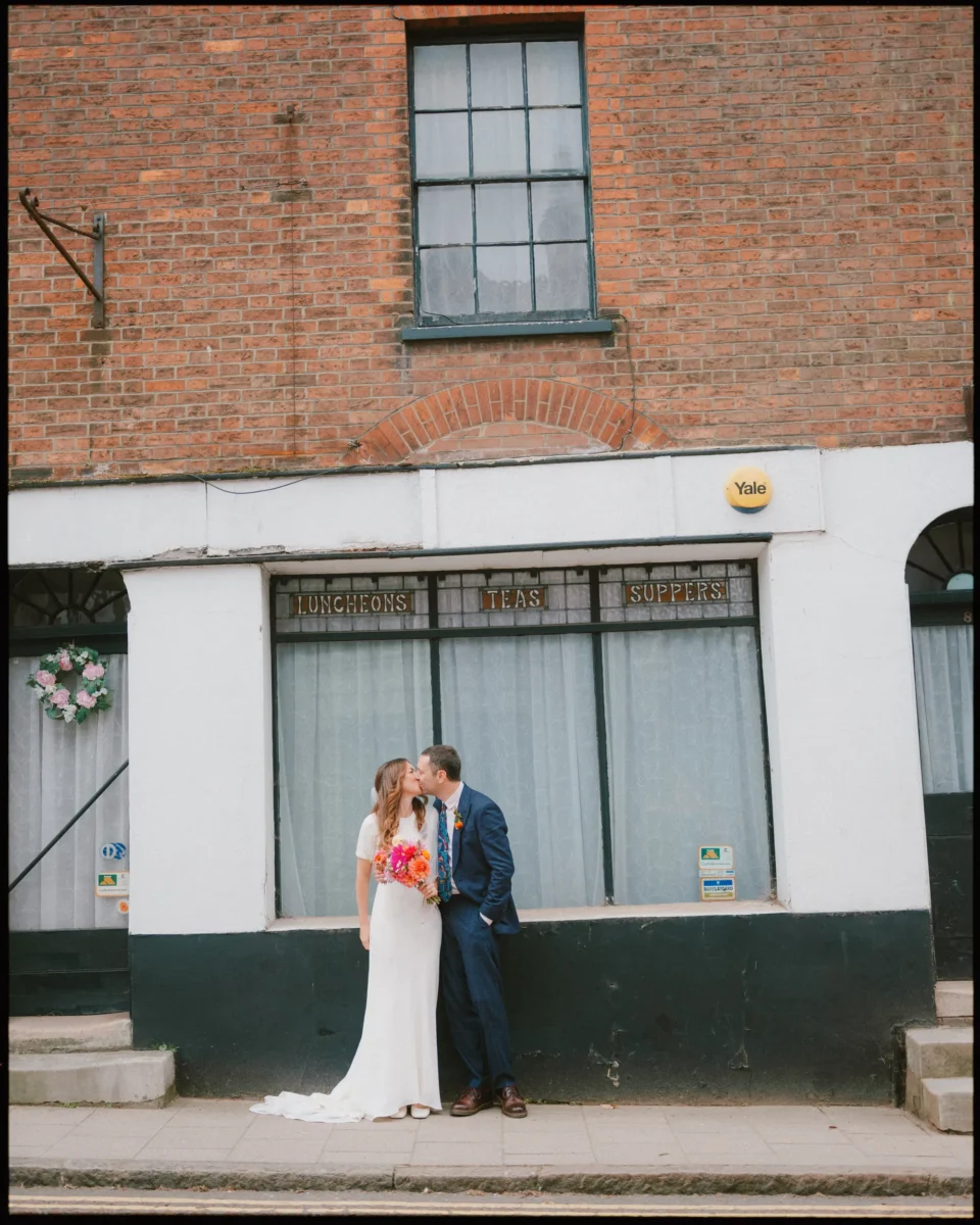 A couple in wedding attire stands on a sidewalk in front of an old brick building with a sign reading Luncheons Teas Suppers. The groom kisses the bride on the forehead while she holds a colorful bouquet.