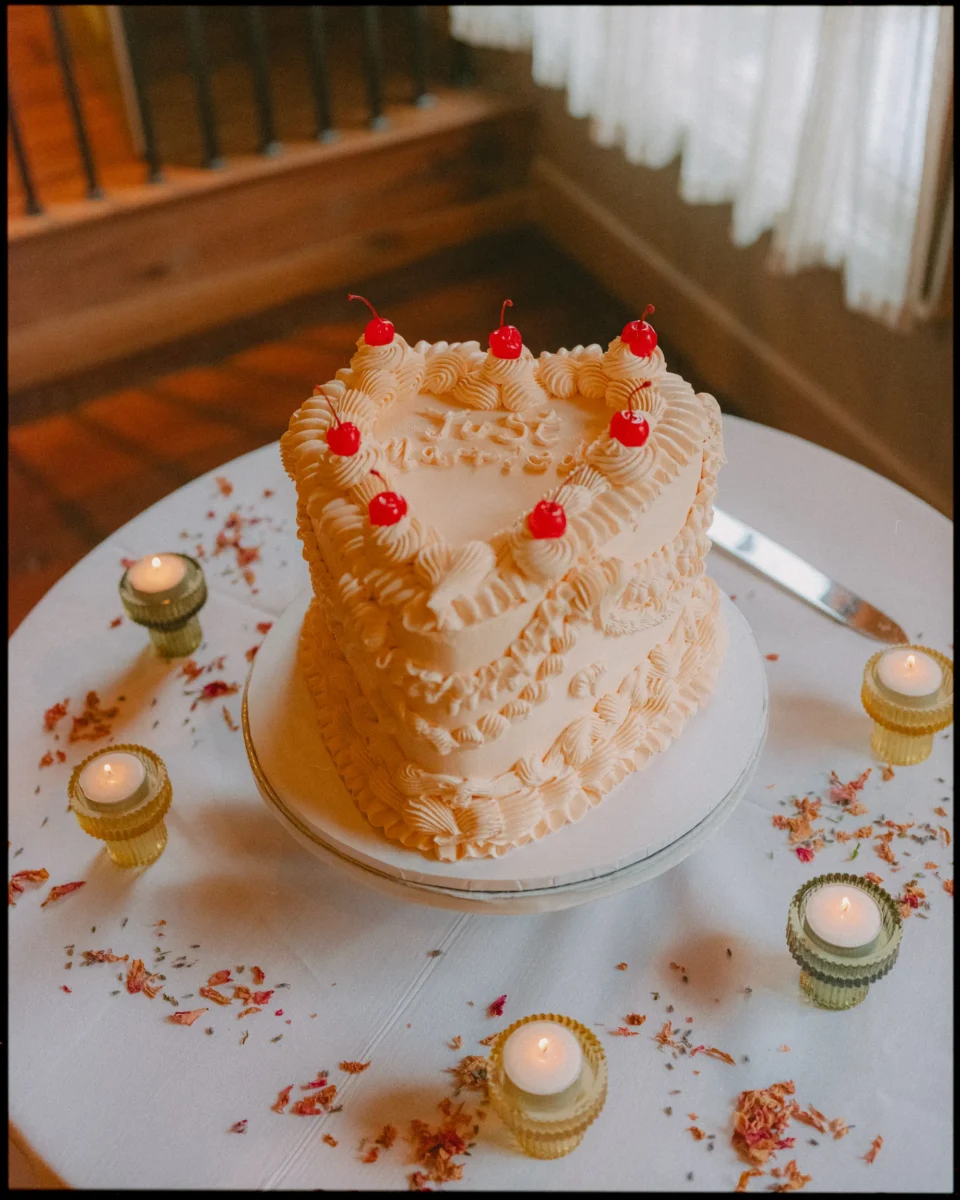 A heart-shaped, two-tiered cake with intricate white frosting and cherries on top sits on a white table surrounded by lit tealight candles and scattered flower petals.