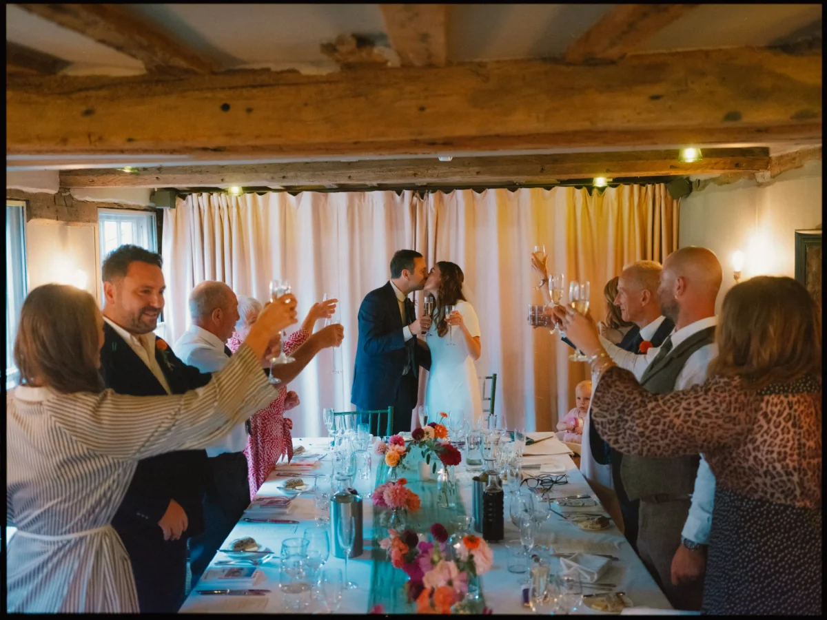 A bride and groom kiss at the head of a long, decorated table as wedding guests stand, raising glasses in a celebratory toast inside a rustic venue with wooden beams and soft lighting.