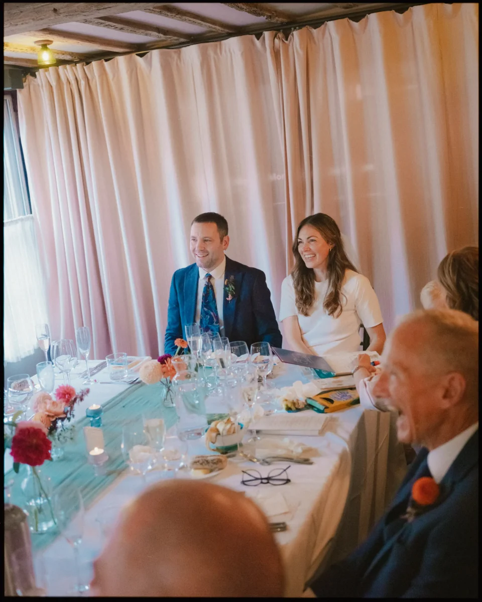 A bride and groom sit smiling at a round table with guests during a wedding reception, surrounded by flowers, candles, and place settings, with soft pink curtains in the background.