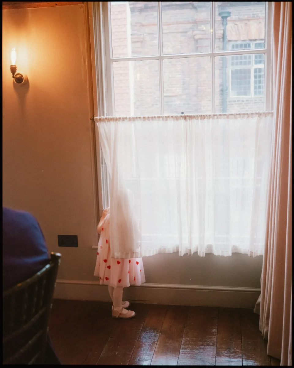 A small child in a white dress with red spots stands on a wooden floor, peeking out from behind a sheer curtain at a tall window, with soft indoor lighting and an old brick building visible outside.