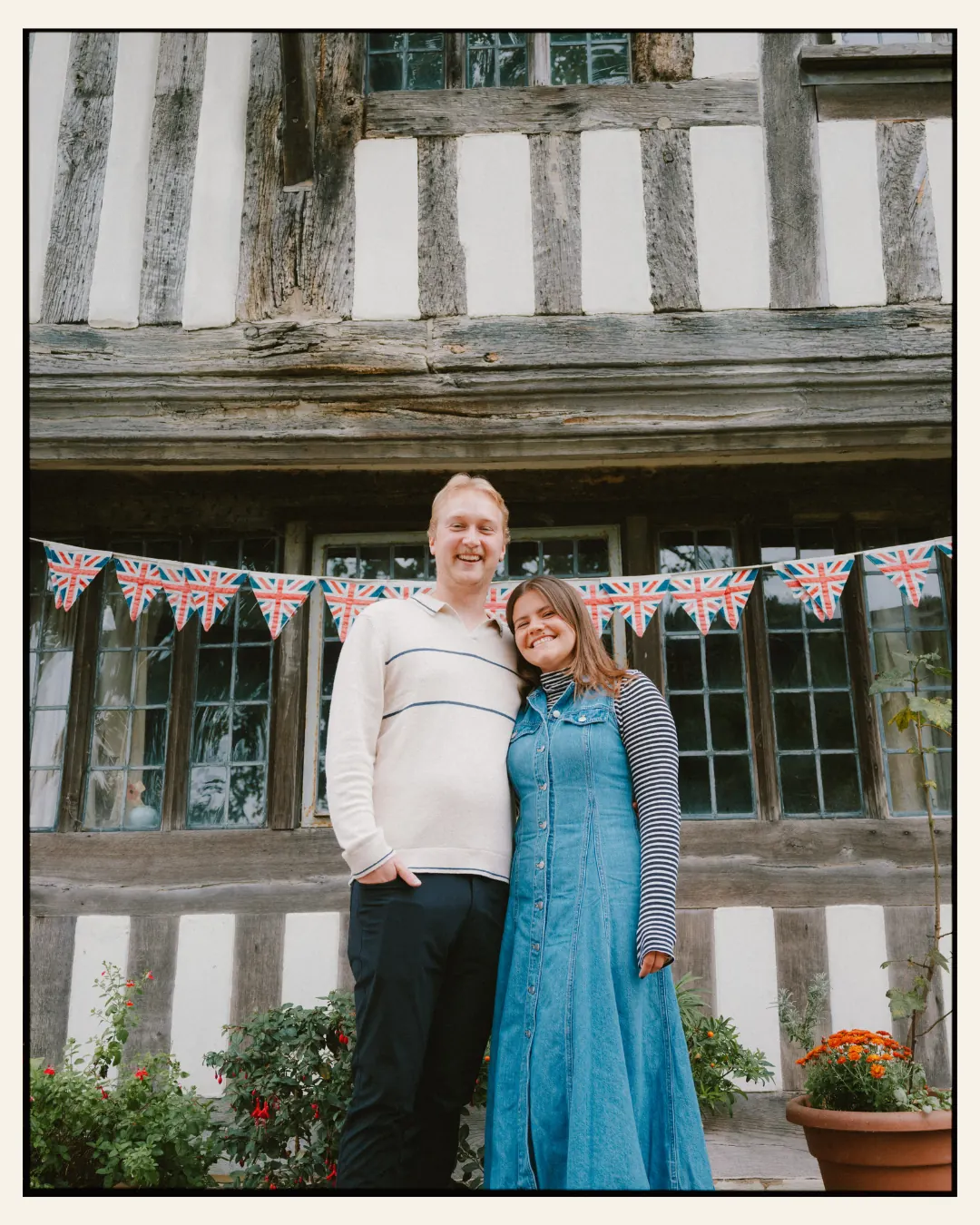 american couple standing in front of union jack bunting on an old tudor building in the ancient town of rye for their engagement shoot ahead of their destination wedding